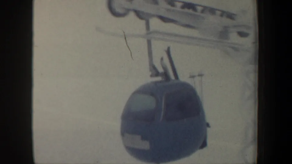 1962 New Hampshire frame showing a blue gondola cable car with skis on top, suspended from overhead cables in a snowy ski-area setting.