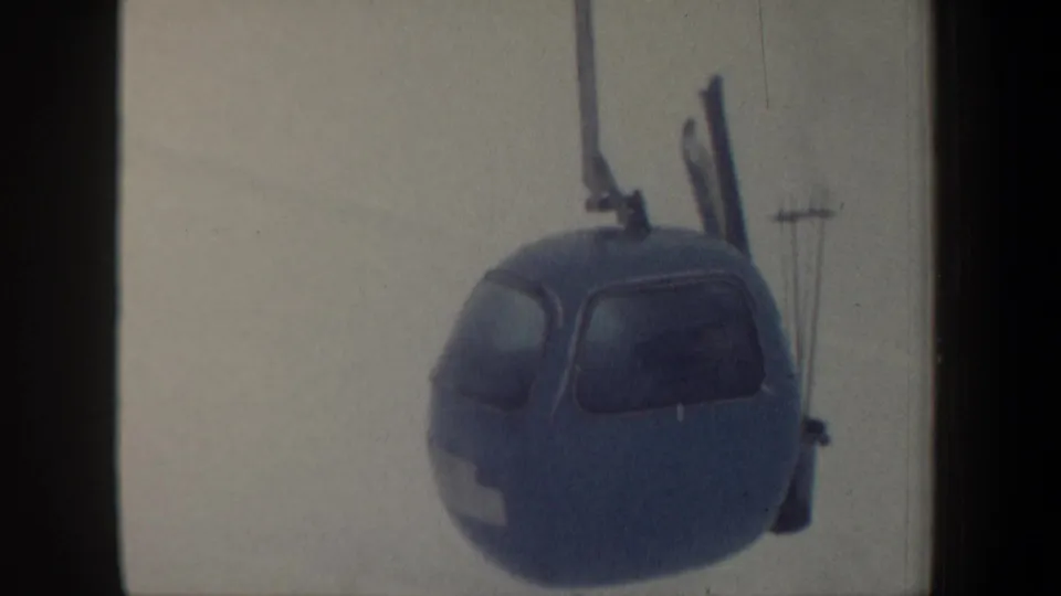 1962 New Hampshire 8mm frame showing a blue gondola lift cabin with exterior skis against a pale snowy mountain backdrop.