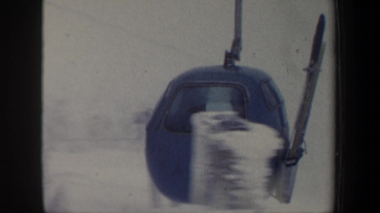 1962 New Hampshire home-movie frame of a blue gondola cabin moving past snowy blur with skis carried on its exterior rack.