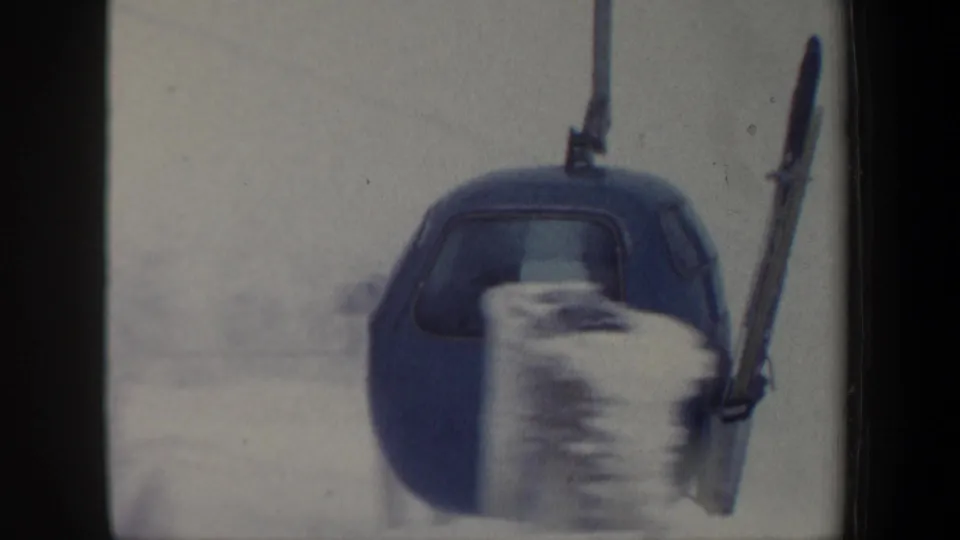 1962 New Hampshire home-movie frame of a blue gondola cabin moving past snowy blur with skis carried on its exterior rack.