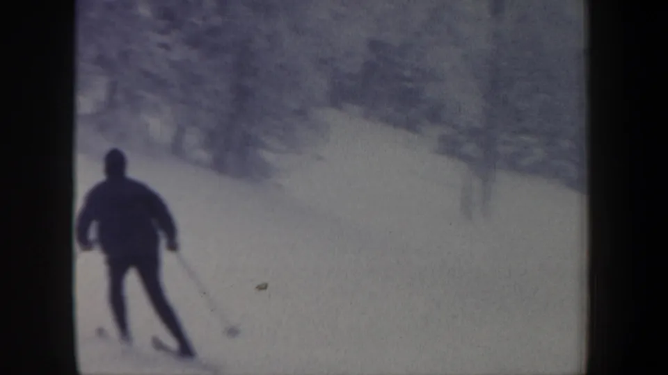 Rear view of an adult skier turning on a snowy wooded slope in 1962 New Hampshire home-movie footage.