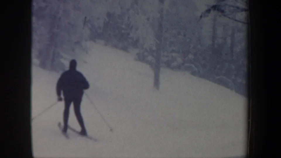 1962 New Hampshire 8mm frame showing a distant skier in dark winter clothing using poles on a snowy wooded slope.