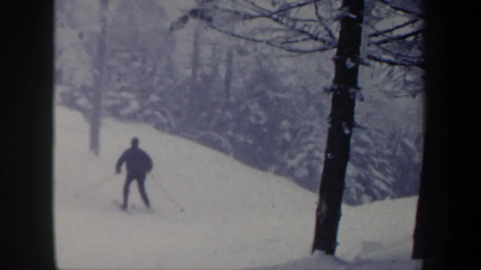 1962 New Hampshire 8mm frame showing a skier from behind descending through a snowy wooded area with poles extended.