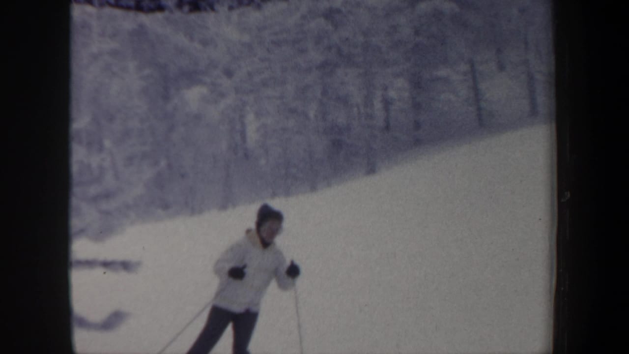 In 1962 New Hampshire home-movie footage, a skier in a light coat, dark pants, knit hat, and gloves descends a snow-covered hill.