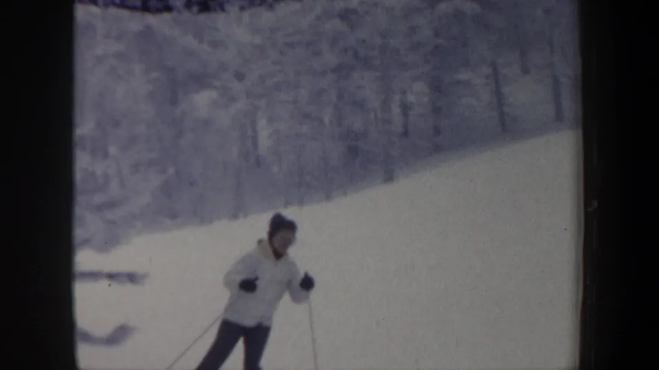 In 1962 New Hampshire home-movie footage, a skier in a light coat, dark pants, knit hat, and gloves descends a snow-covered hill.