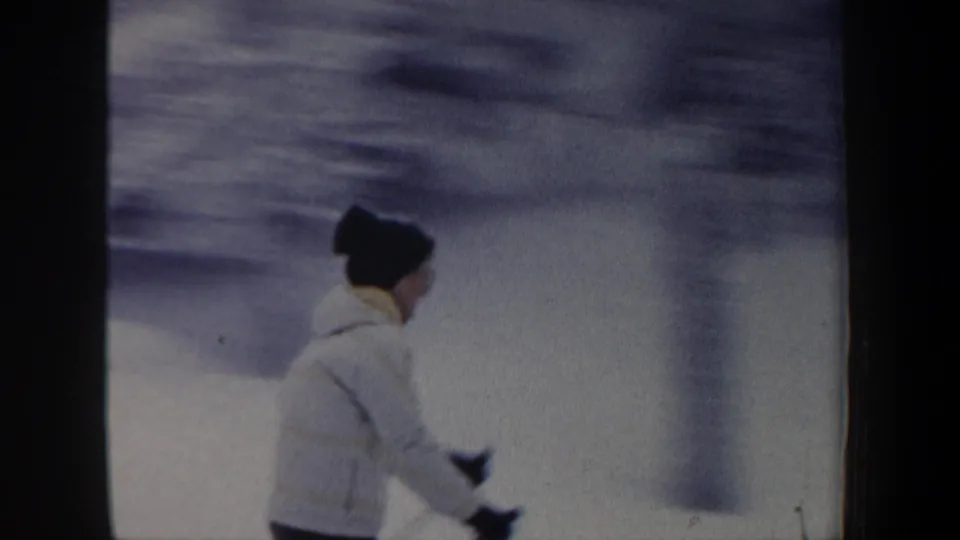 The skier passes in profile across the snowy New Hampshire slope, wearing a light jacket and dark hat while holding ski poles.