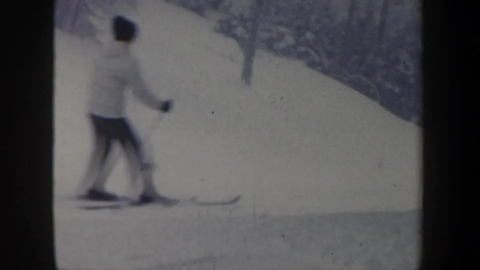 A softly blurred home-movie frame shows the skier moving across the snow, with bare winter trees rising beyond the slope.