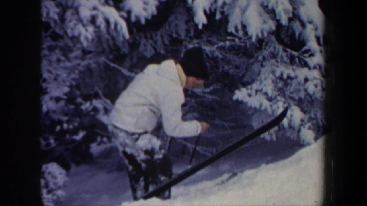 Skier in a white jacket bends beside snow-covered evergreens on a snowy slope.