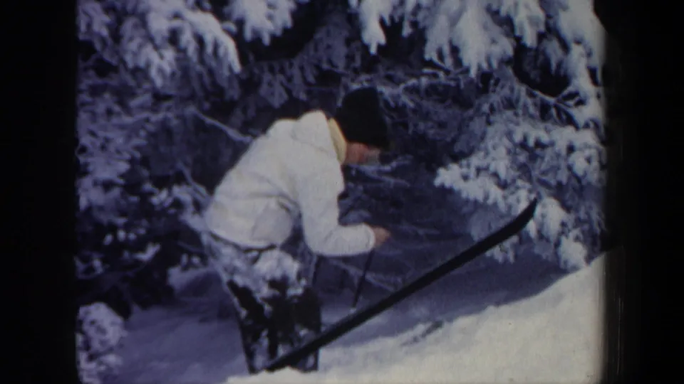 1962 New Hampshire 8mm frame of a skier in a white jacket leaning near deep snow beneath snow-laden evergreen branches.