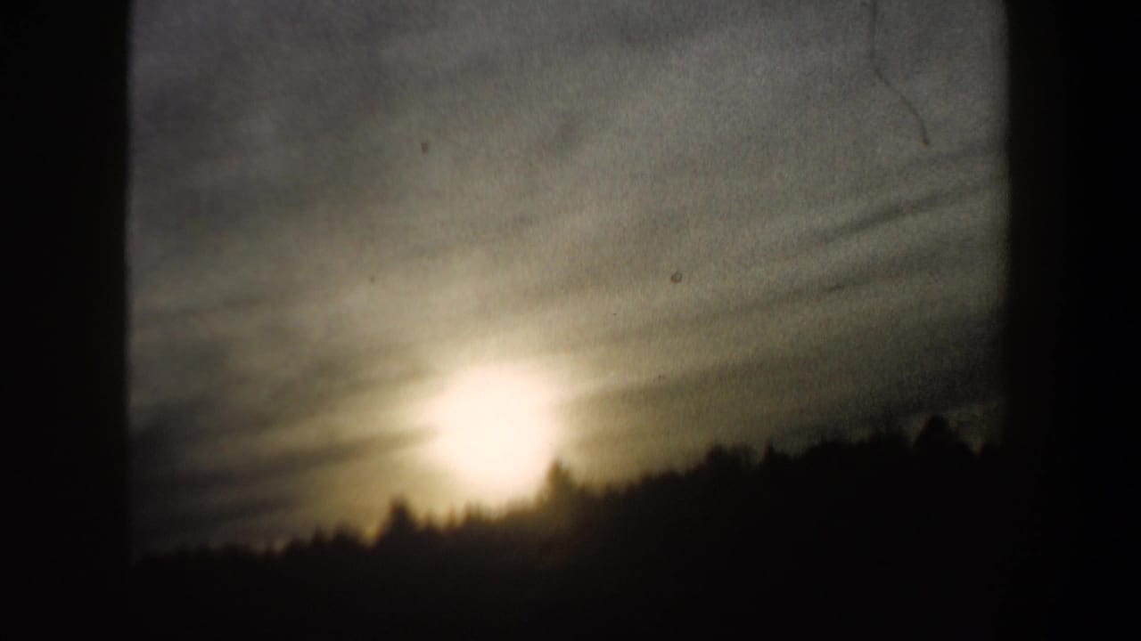 1962 New Hampshire home-movie frame showing strong low light over a winter treeline, with soft focus and dark 8mm frame edges.
