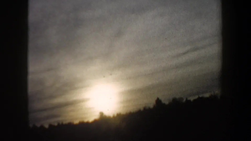 1962 New Hampshire 8mm frame with a dim sky, low winter glare, and a dark treeline, seen through soft grain and exposure flare.