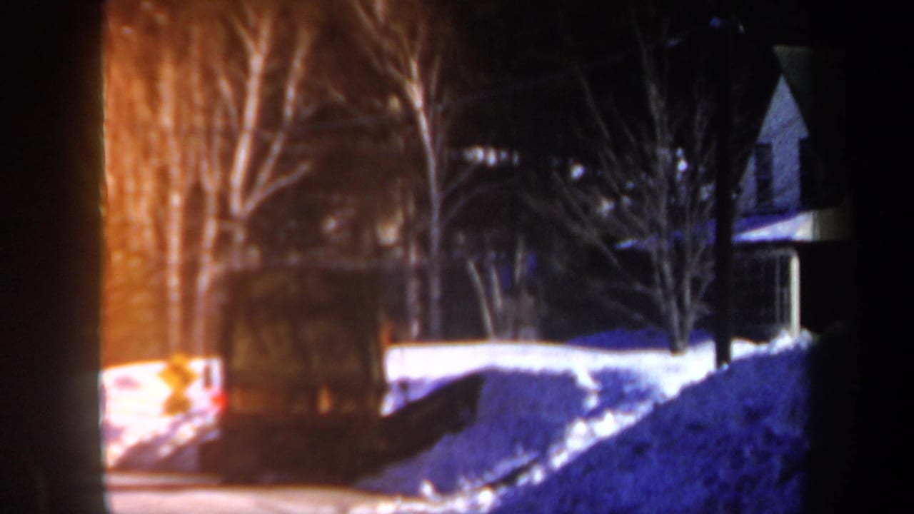 Snowy house exterior at night with bare trees and a partial vehicle at the frame edge.