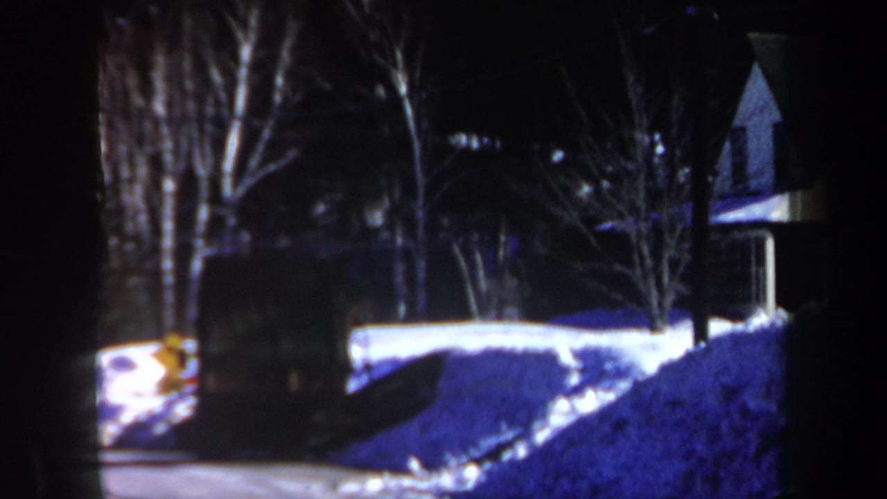 1962 New Hampshire home-movie frame of snow piled along a residential property, with bare trees and a house partly visible in low light.