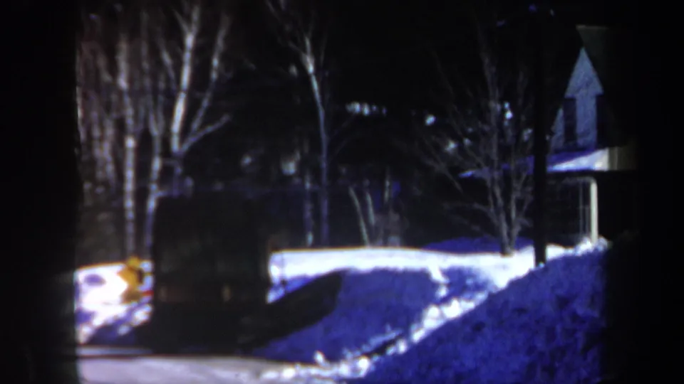 1962 New Hampshire home-movie frame of snow piled along a residential property, with bare trees and a house partly visible in low light.