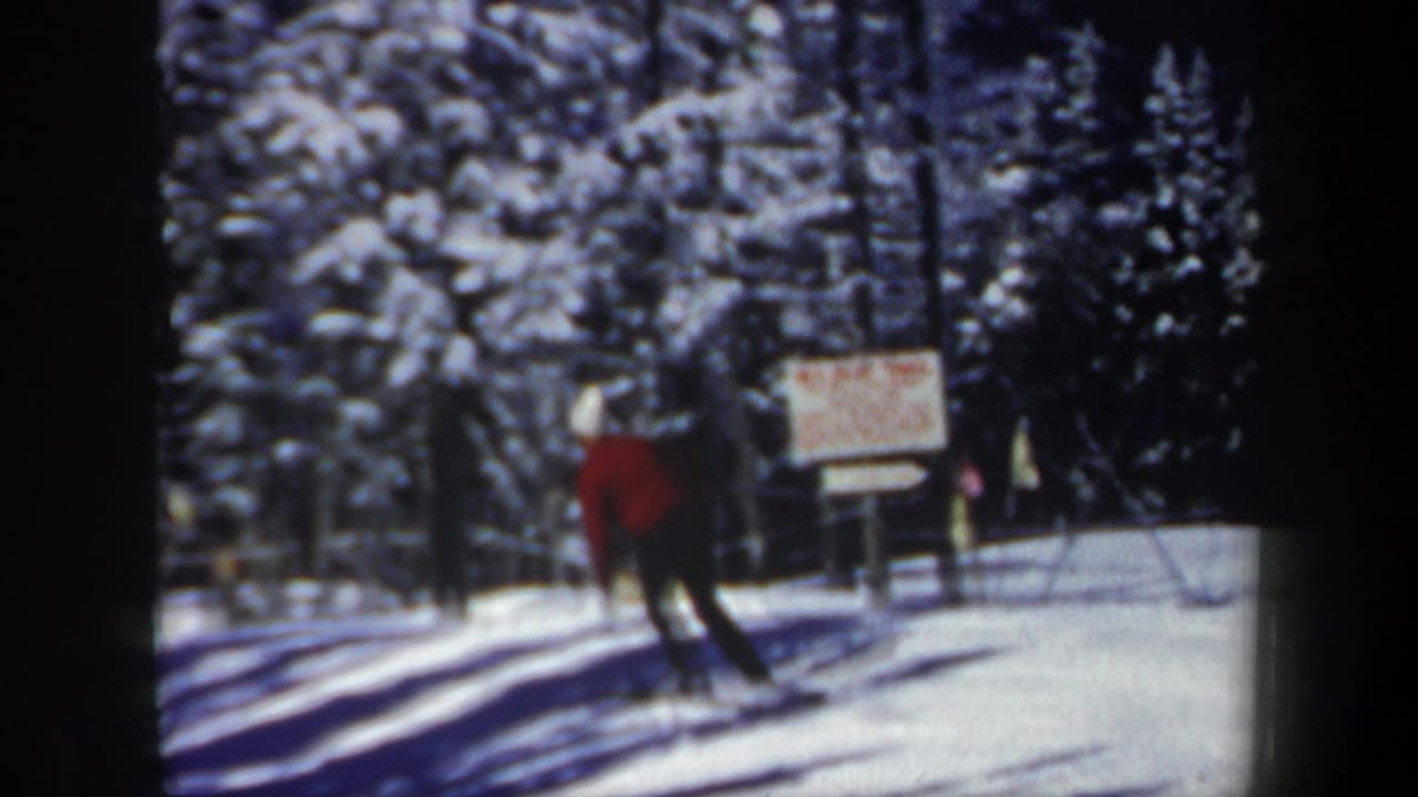 1962 New Hampshire home-movie frame showing a winter-clad man gliding near a trail sign in a snowy evergreen area.