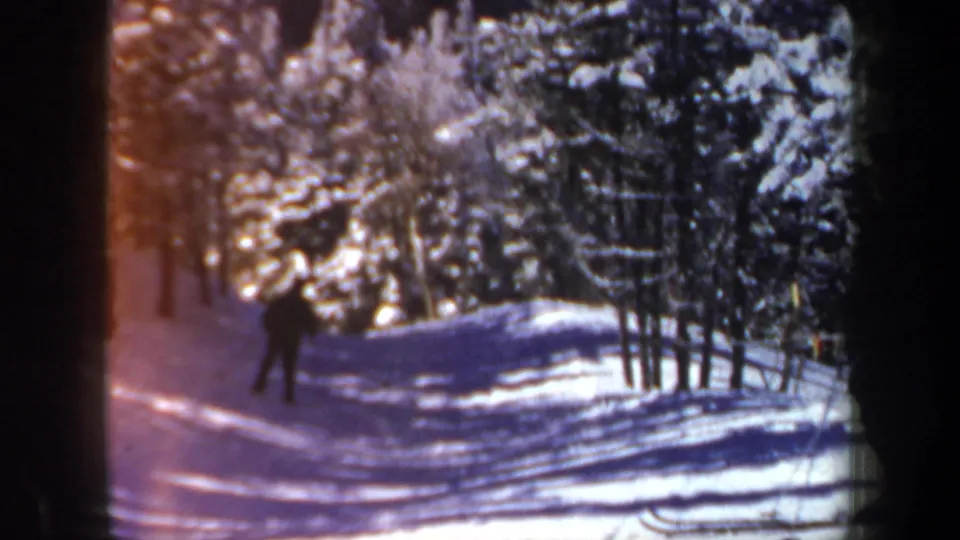 1962 New Hampshire home-movie frame with a skier farther down a snow-covered forest path in blue shadow.