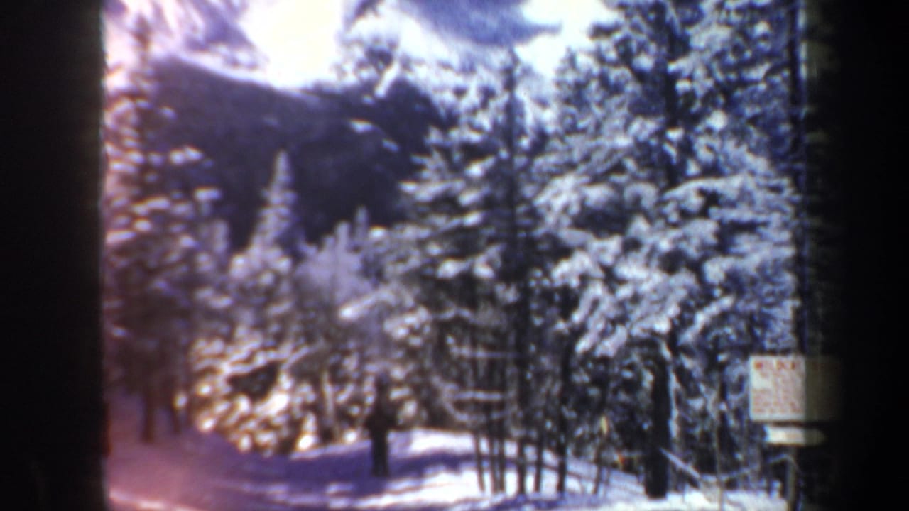 1962 New Hampshire home-movie frame showing a distant skier in a snowy evergreen setting with a small posted sign and warm film flare.
