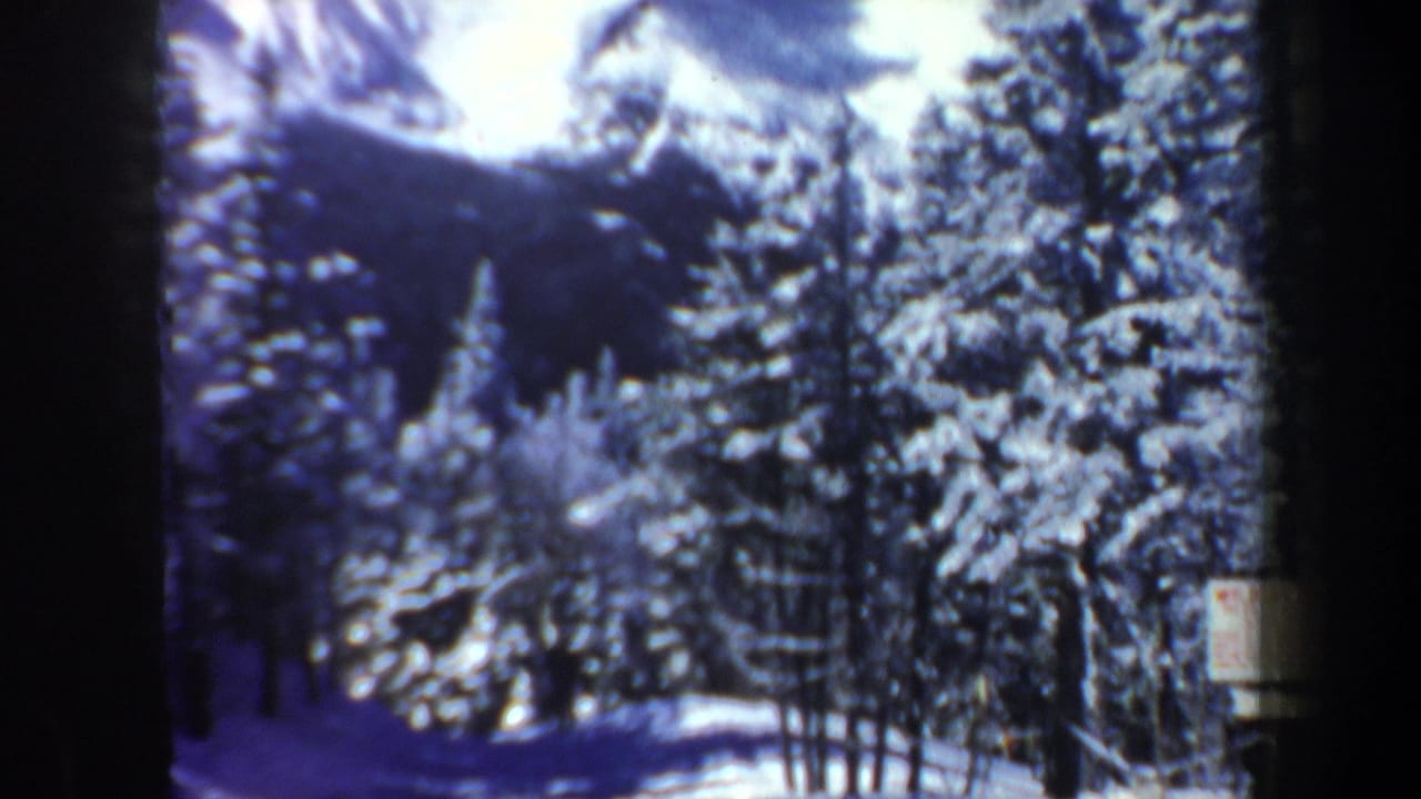 1962 New Hampshire 8mm frame showing snowy evergreens, distant mountain shapes, and a small sign near a winter slope.