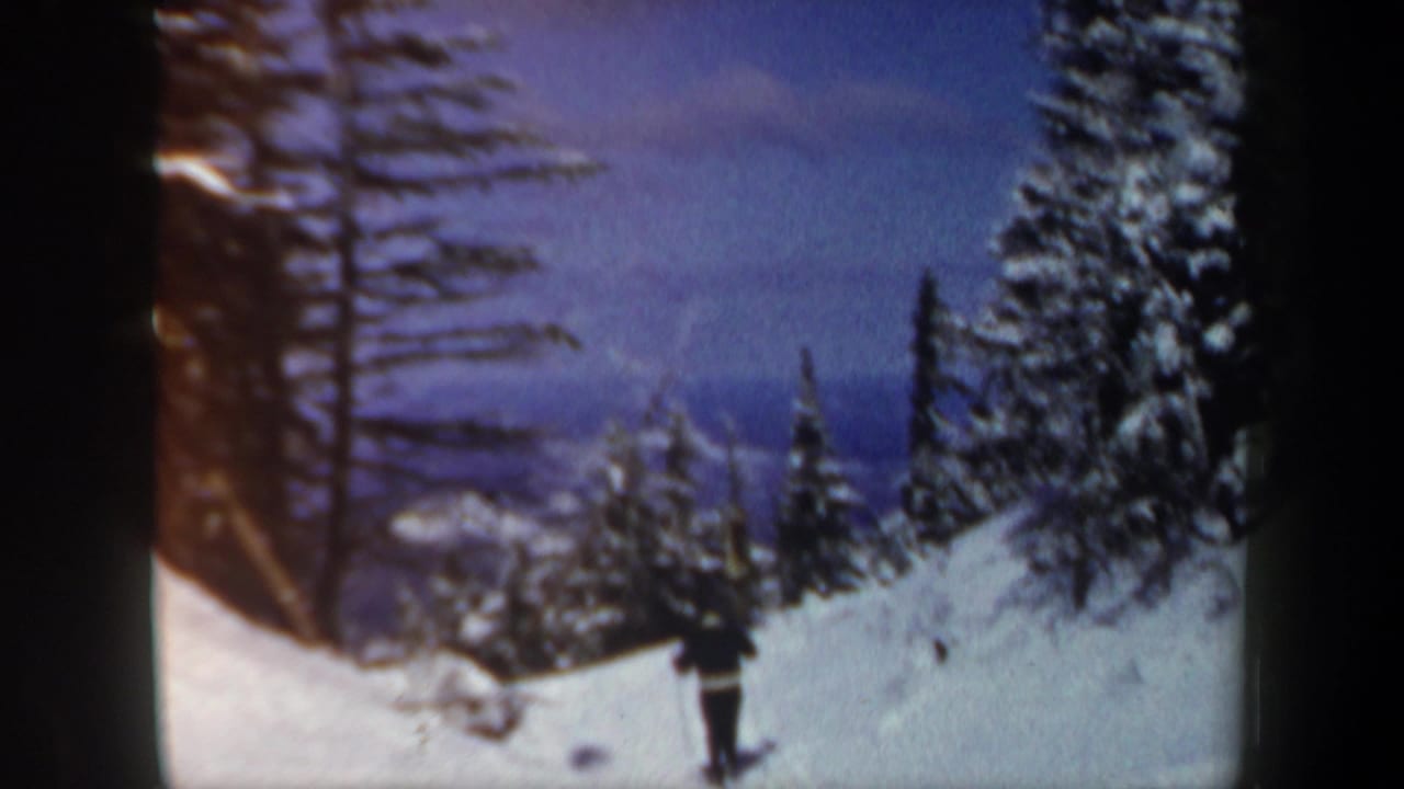 1962 New Hampshire 8mm frame showing a distant lone skier moving along a snowy evergreen-lined mountain slope.