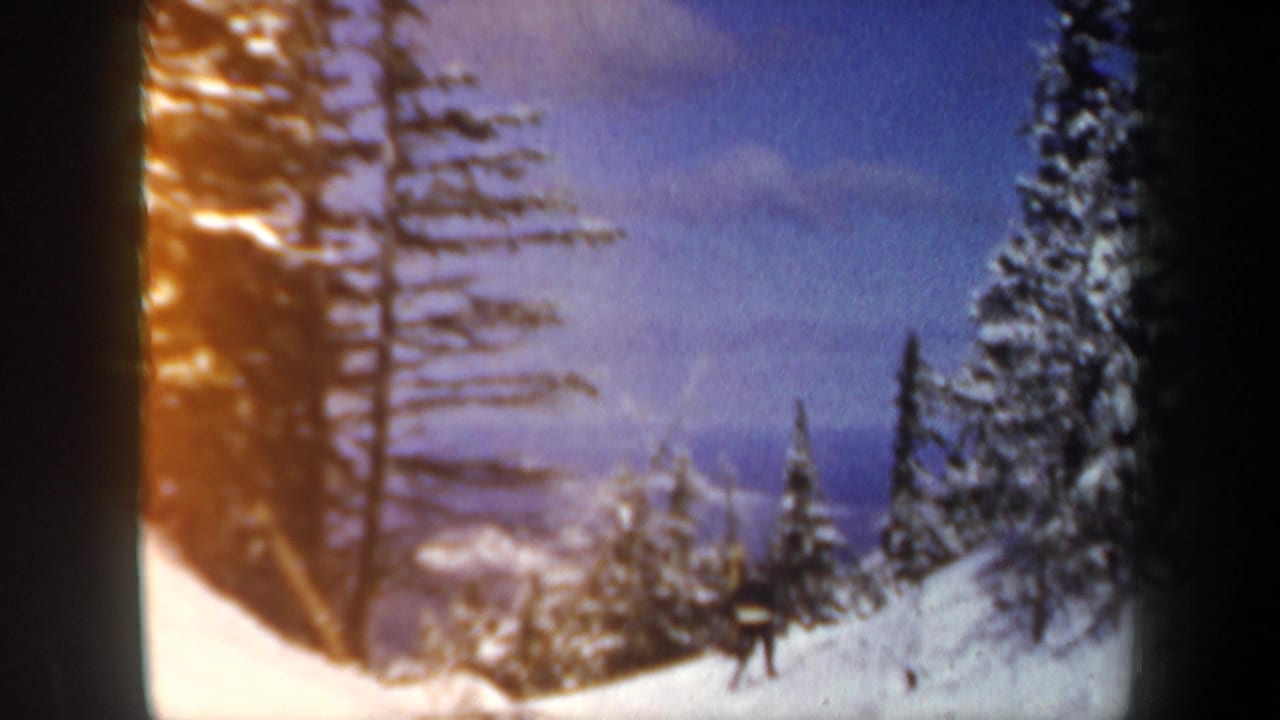1962 New Hampshire home-movie frame of a small skier figure crossing a bright snowy slope among conifers.