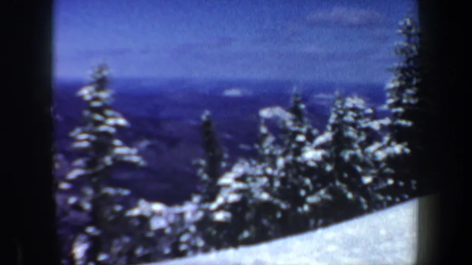 1962 New Hampshire 8mm frame showing a snowy foreground slope, clustered evergreens, and hazy blue ridges in the distance.