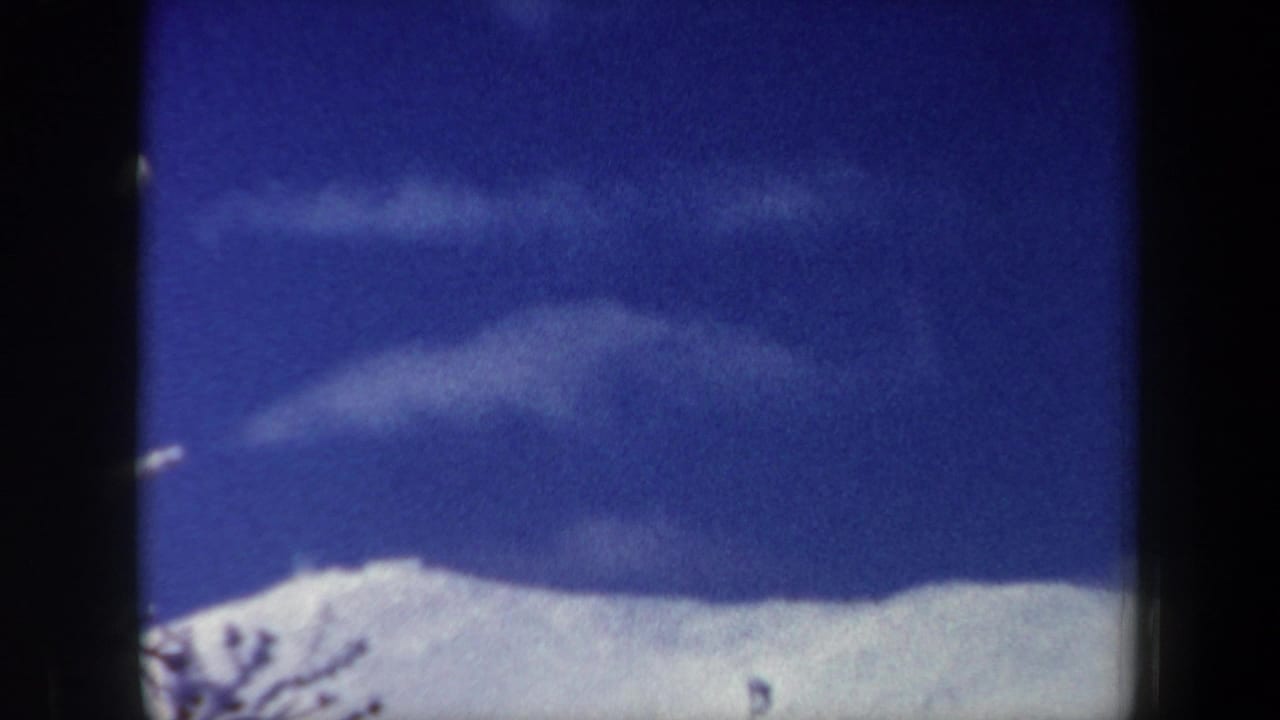 1962 New Hampshire home-movie frame with a snowy ridge, deep blue sky, and pale clouds, showing strong 8mm grain and gate edges.