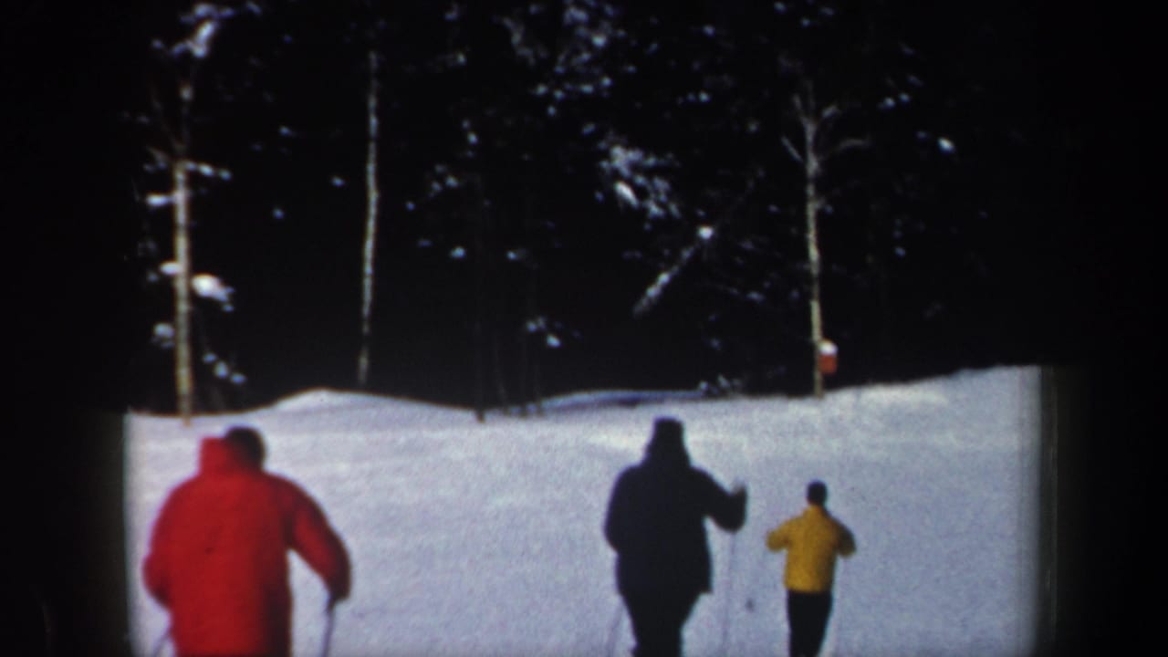 1962 New Hampshire home-movie frame showing three distant skiers descending a snowy hill with dark trees and utility poles beyond.