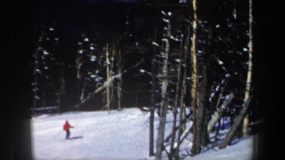 1962 New Hampshire home-movie frame showing a distant skier in red crossing a bright snowy slope near dark pines.