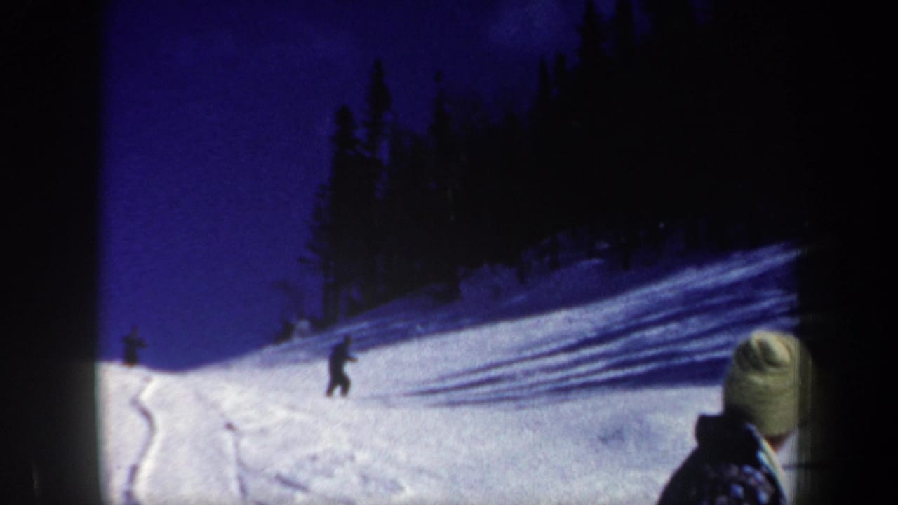 Skiers descend a snowy slope while a bundled viewer watches from the foreground.