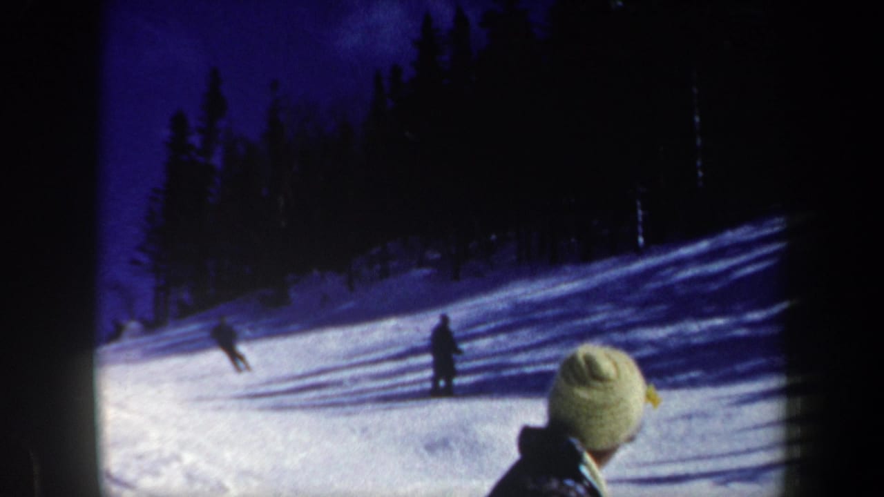 Two skiers cross a snowy hillside near a foreground spectator in a knit hat.