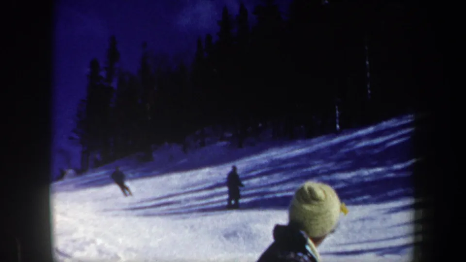 Blue-toned 1962 New Hampshire ski-slope frame with two distant skiers and a bundled foreground viewer.