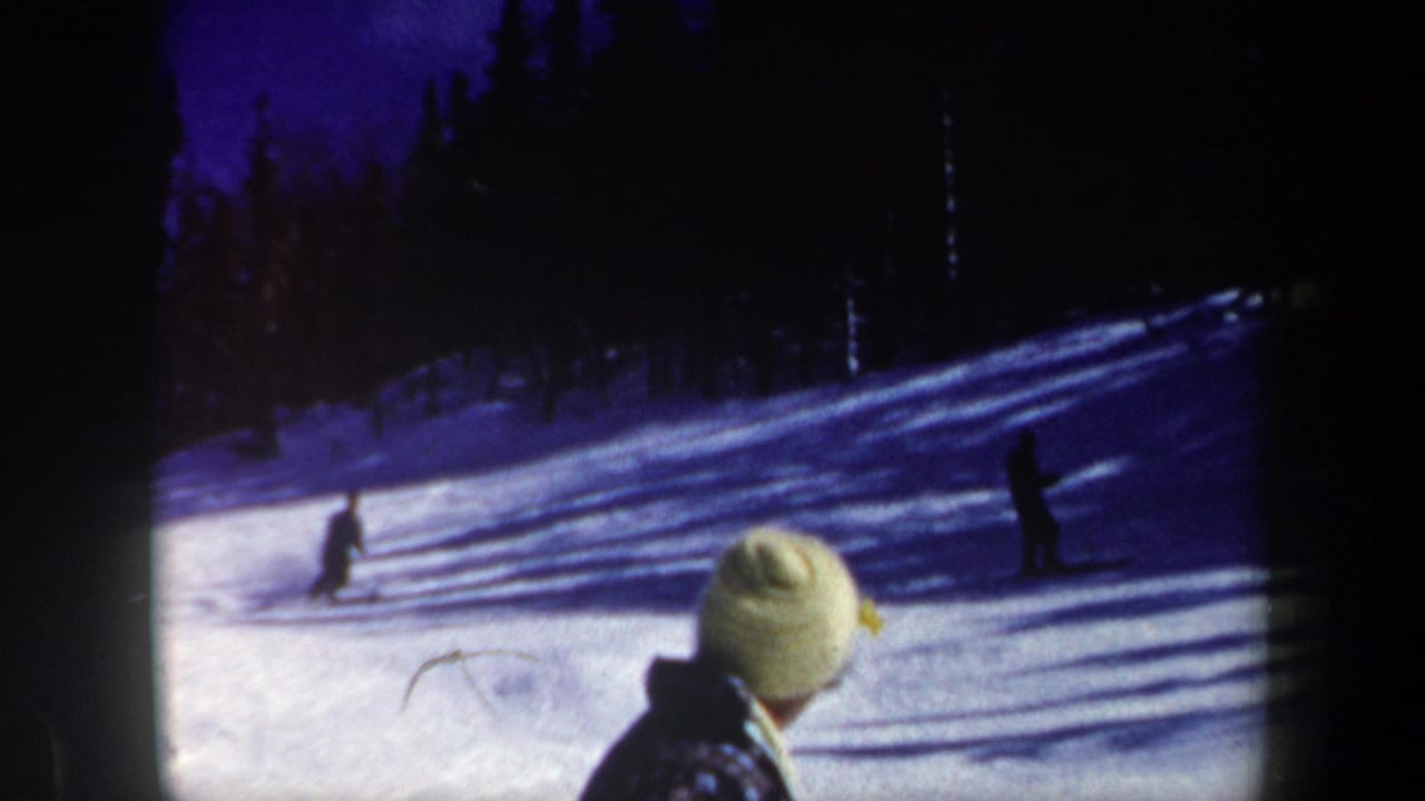 1962 New Hampshire home-movie frame showing distant skiers, evergreen trees, and a spectator in a light knit hat.