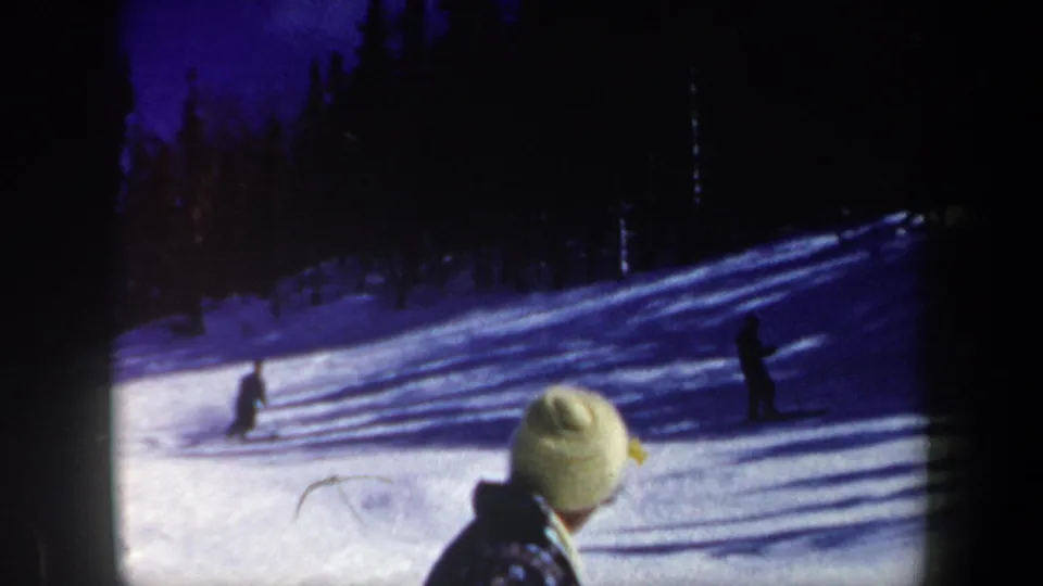 1962 New Hampshire home-movie frame showing distant skiers, evergreen trees, and a spectator in a light knit hat.