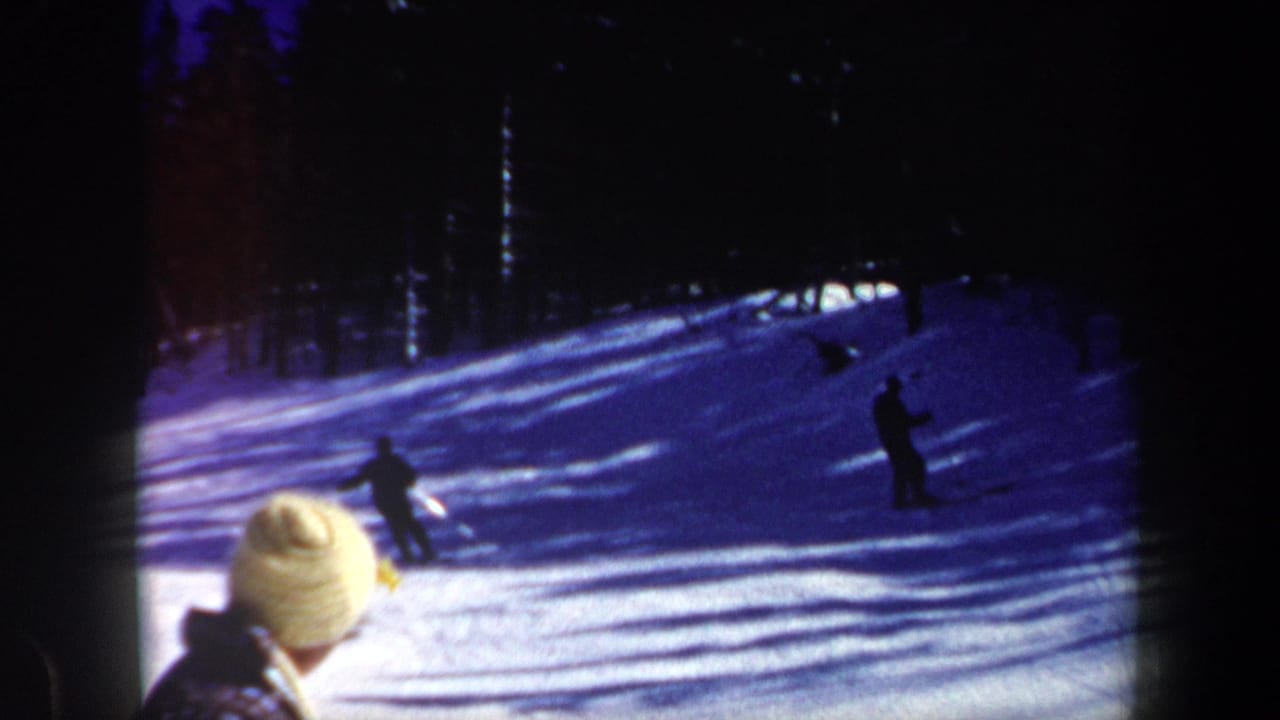 1962 New Hampshire 8mm frame with skiers on a shaded snowy hillside and a bundled viewer in the foreground.
