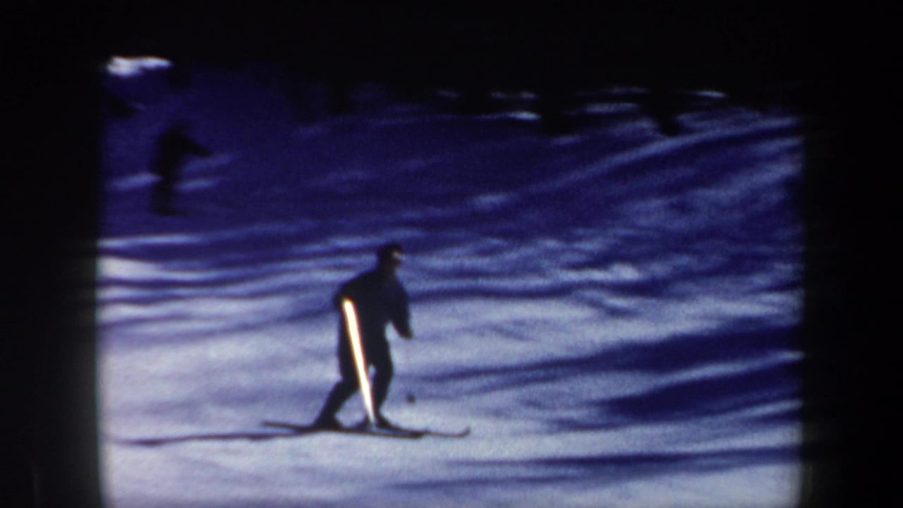 A dark-clad skier crosses a snowy slope with trees and another distant skier behind.