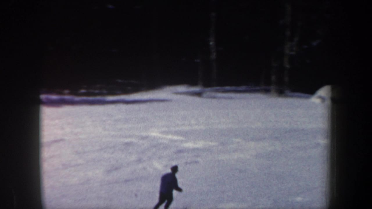 1962 New Hampshire 8mm frame with a small dark skier figure crossing a wide pale snowfield beneath a shadowed tree line.