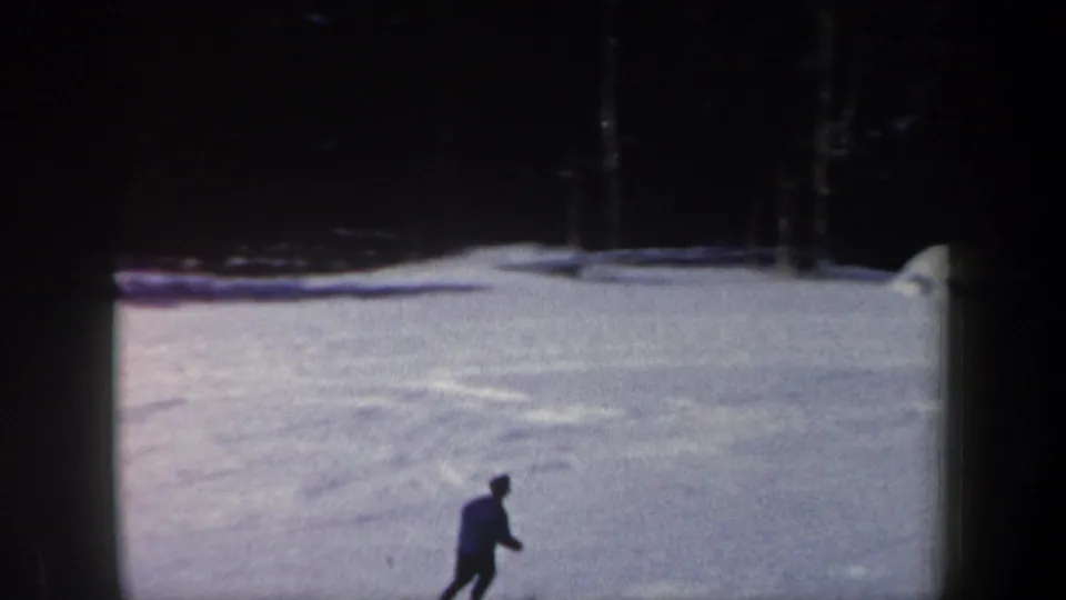 1962 New Hampshire 8mm frame with a small dark skier figure crossing a wide pale snowfield beneath a shadowed tree line.