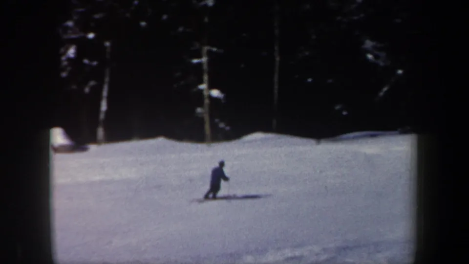 1962 New Hampshire 8mm frame showing a skier moving across an open slope bordered by dark evergreens and strong winter shadows.