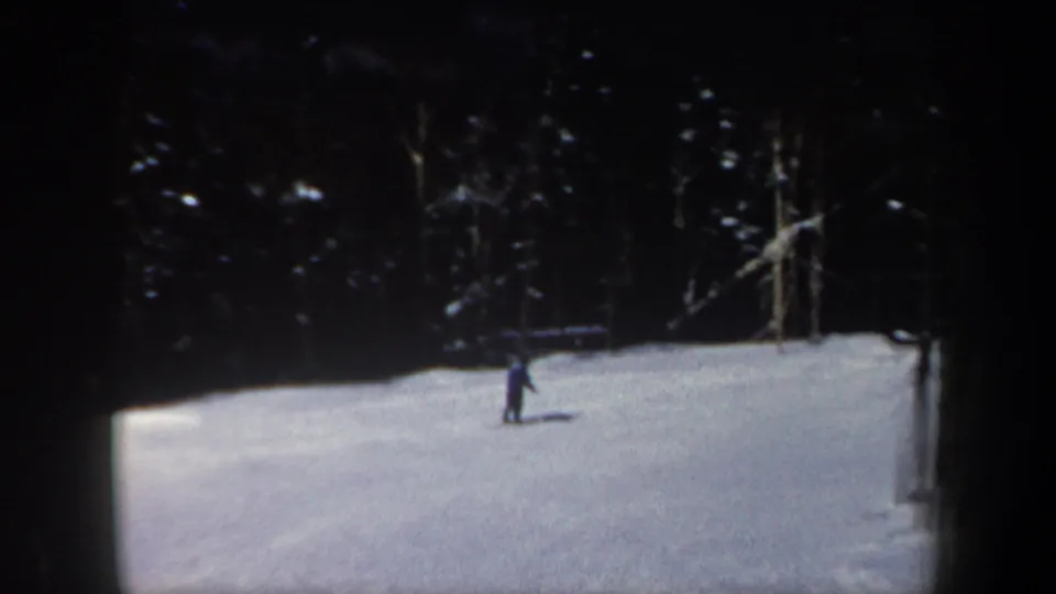 1962 New Hampshire home-movie frame with a lone skier on a sunlit slope backed by dense evergreens.