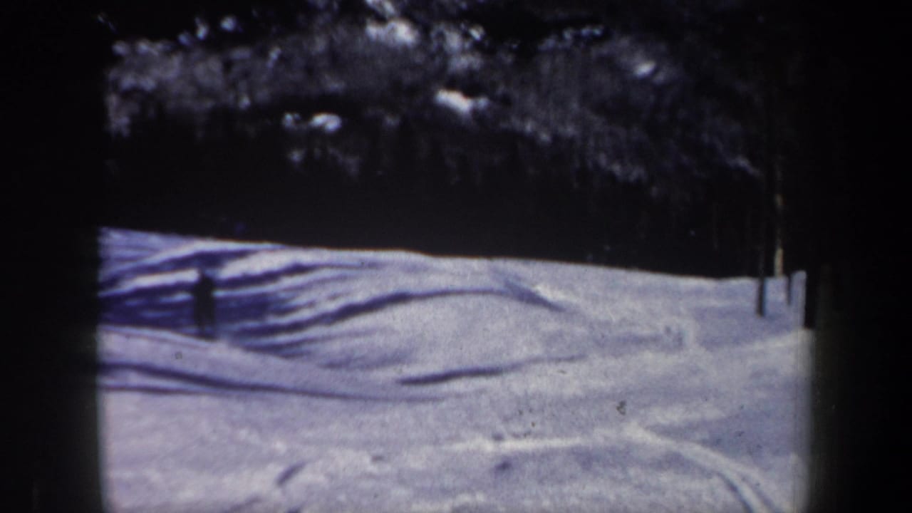 1962 New Hampshire 8mm frame showing a quiet snowy slope, evergreen tree line, visible ski tracks, and strong winter shadows.
