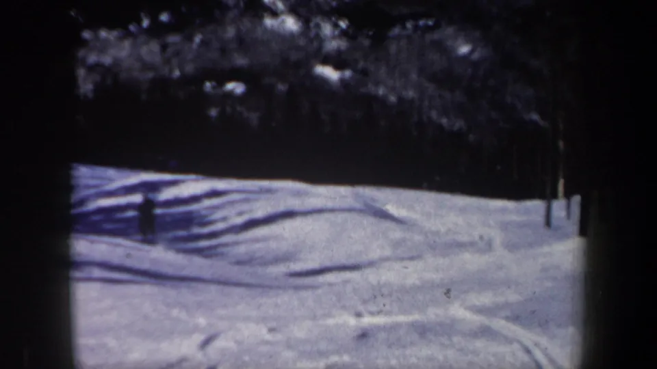 1962 New Hampshire 8mm frame showing a quiet snowy slope, evergreen tree line, visible ski tracks, and strong winter shadows.