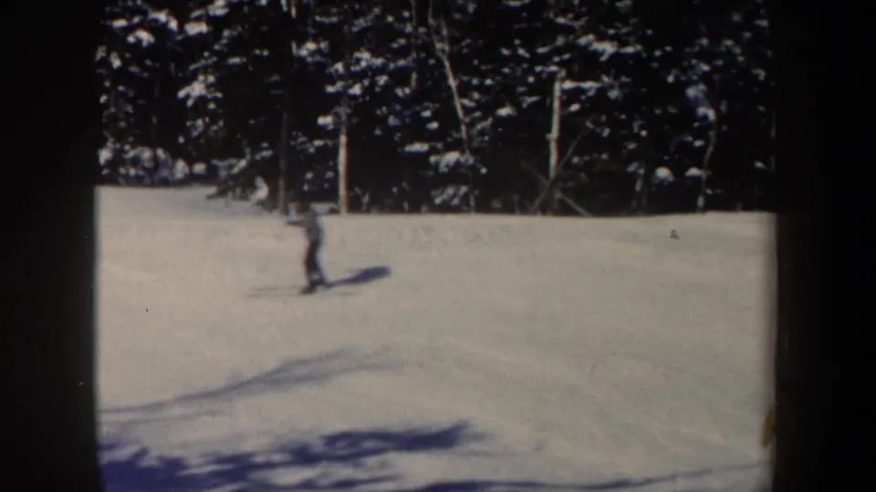 1962 New Hampshire ski-slope frame showing a distant skier with poles, snowy terrain, tree line, and vertical posts in the background.