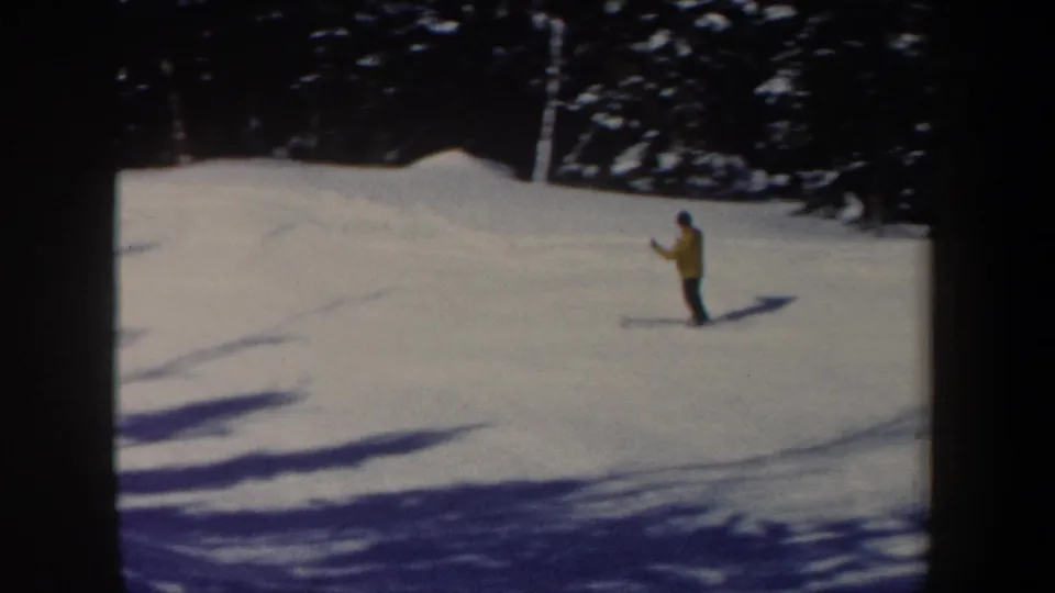 1962 New Hampshire home-movie frame with a yellow-jacketed skier on open snow below a wooded edge.