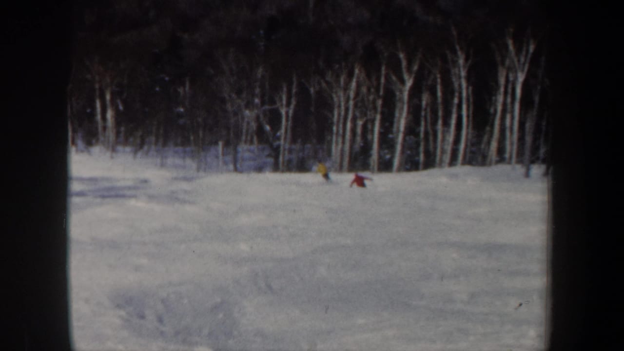 1962 New Hampshire 8mm frame showing two distant skiers on a white slope bordered by leafless woodland.