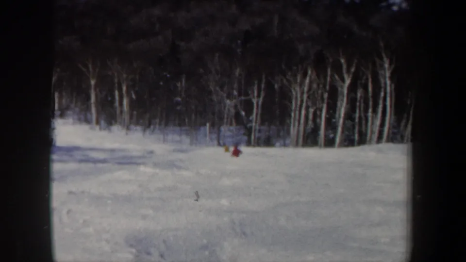 A 1962 New Hampshire home-movie frame shows red and yellow jacketed skiers on a wide snowy hillside near bare trees.