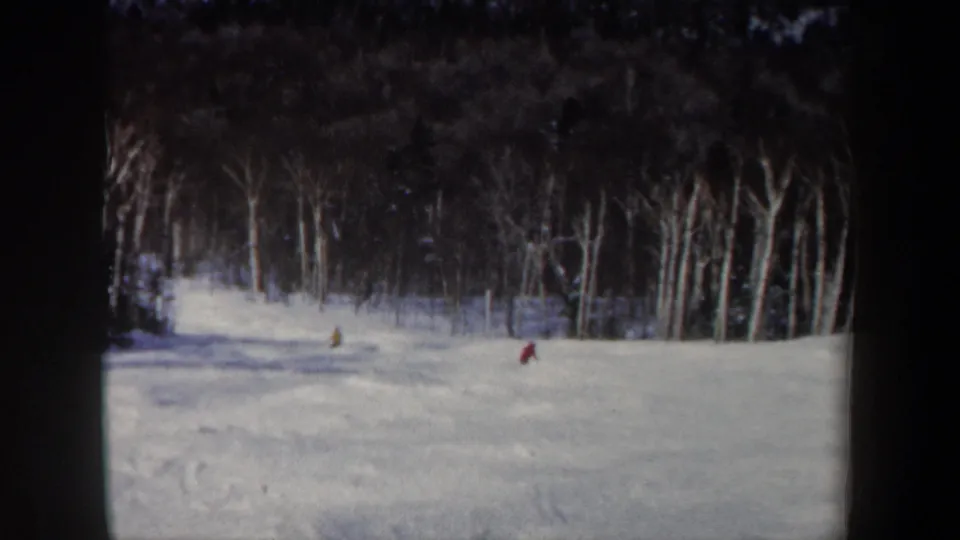 1962 New Hampshire 8mm footage frame with two small skiers separated across a broad snowy slope below bare woodland.