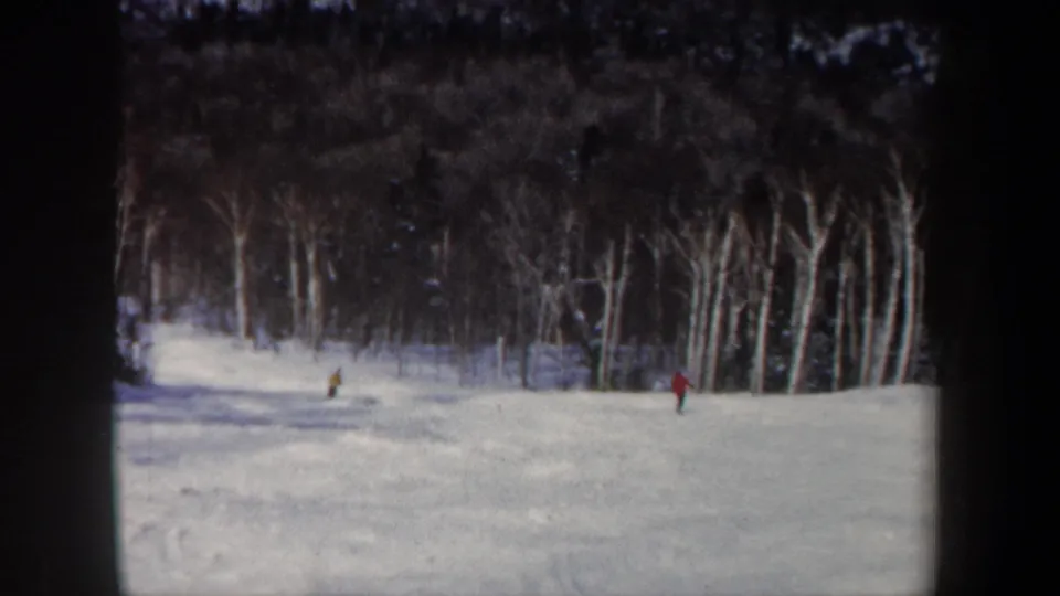 Final 1962 New Hampshire 8mm frame showing two distant skiers on a pale snow slope with bare trees behind them.