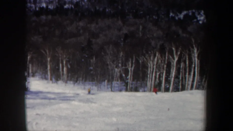 1962 New Hampshire 8mm frame showing distant skiers on a snow-covered slope at the edge of a leafless forest.