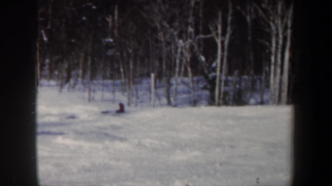 A 1962 New Hampshire home-movie frame shows a distant skier sitting or crouching on a snowy slope beside bare trees.