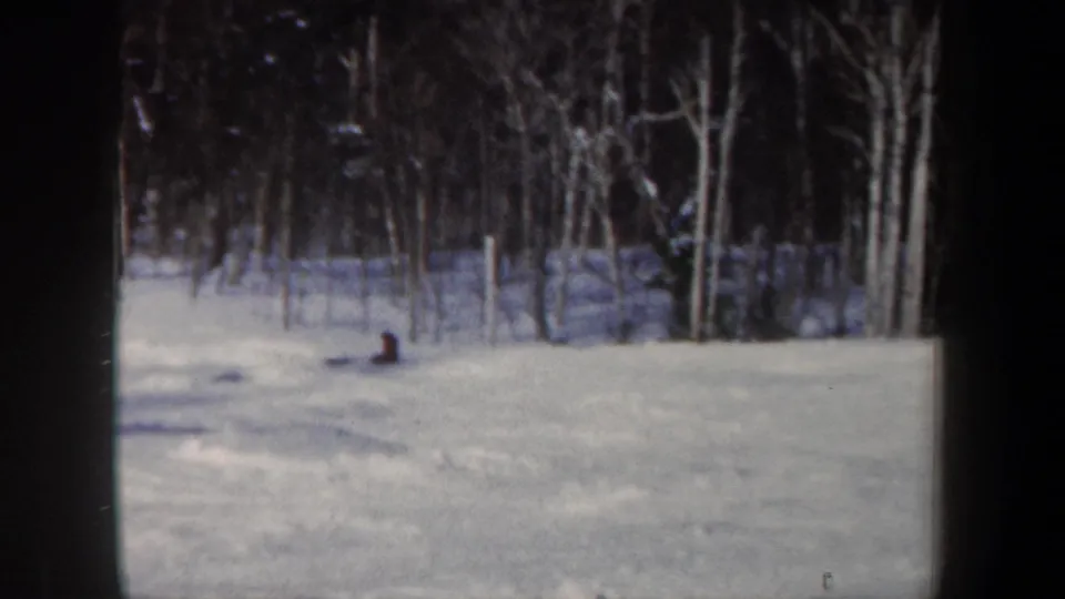 A 1962 New Hampshire home-movie frame shows a distant skier sitting or crouching on a snowy slope beside bare trees.